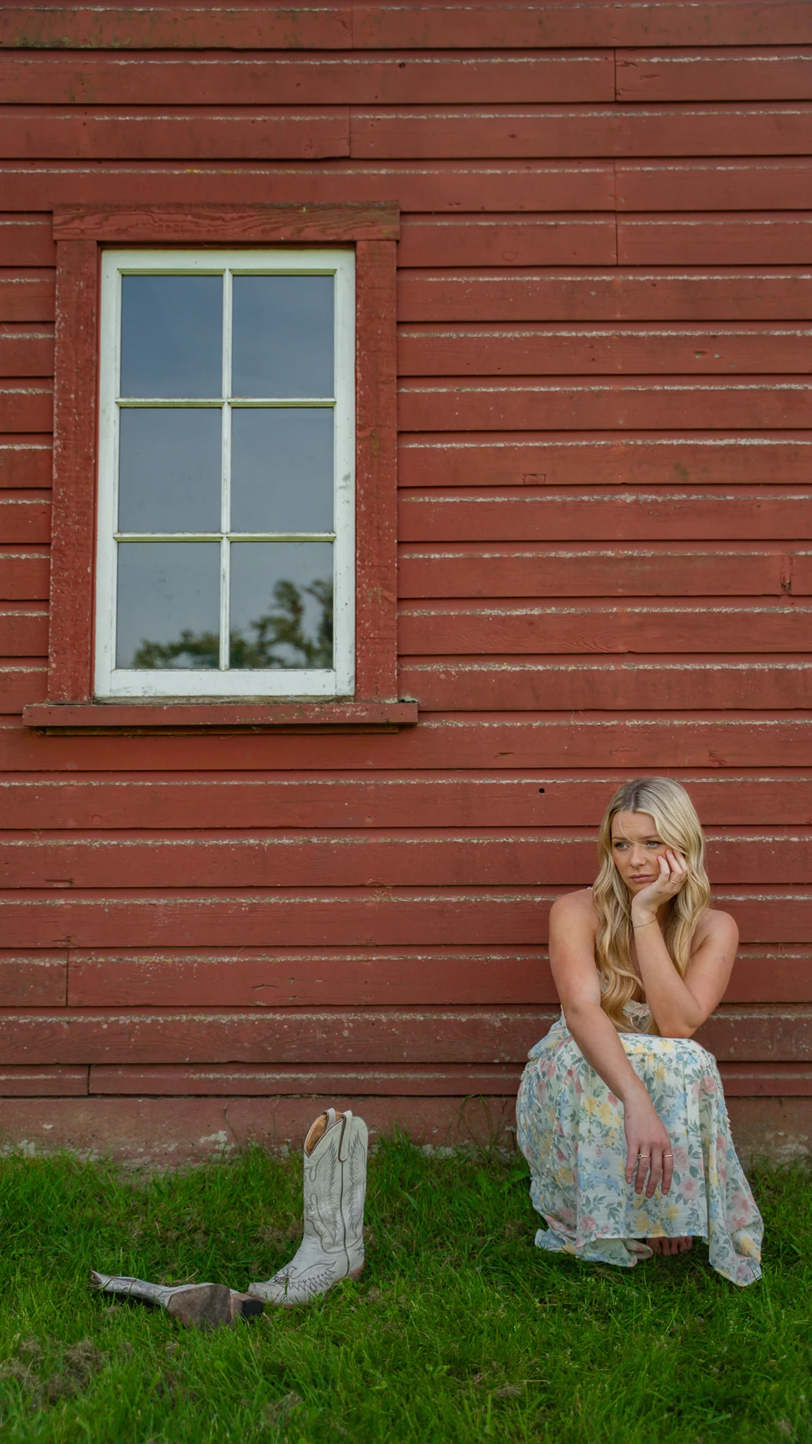Emily Nelson sitting beside a weathered red barn with her cowboy boots in the grass